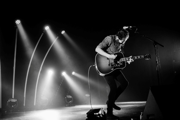 Black and white photo of musician Passenger on stage with stage lights behind.